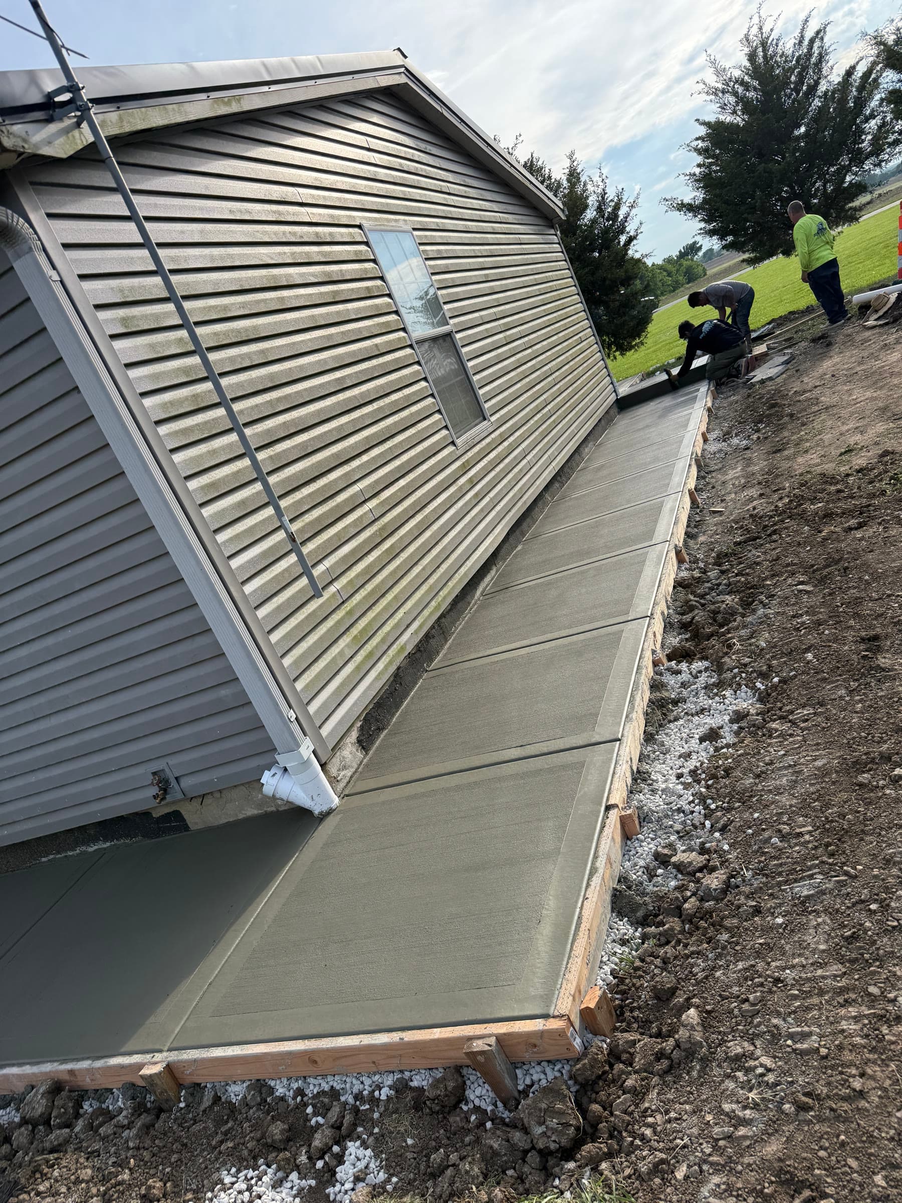 Freshly poured concrete retaining wall along a graded slope at a rural property, crew members visible in the background