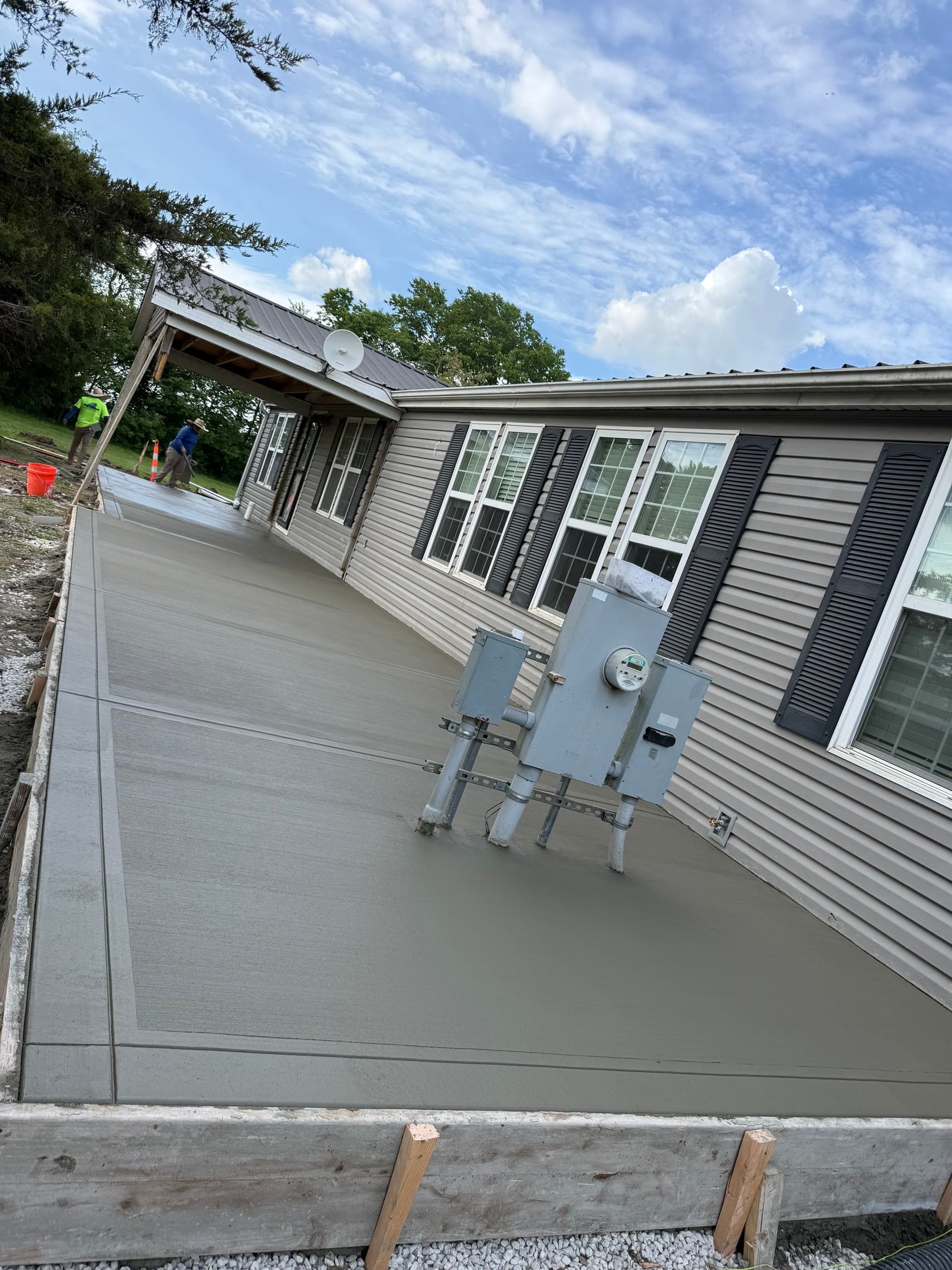 Finished broom-finished concrete patio slab at a rural property, electrical meter boxes visible on the exterior wall