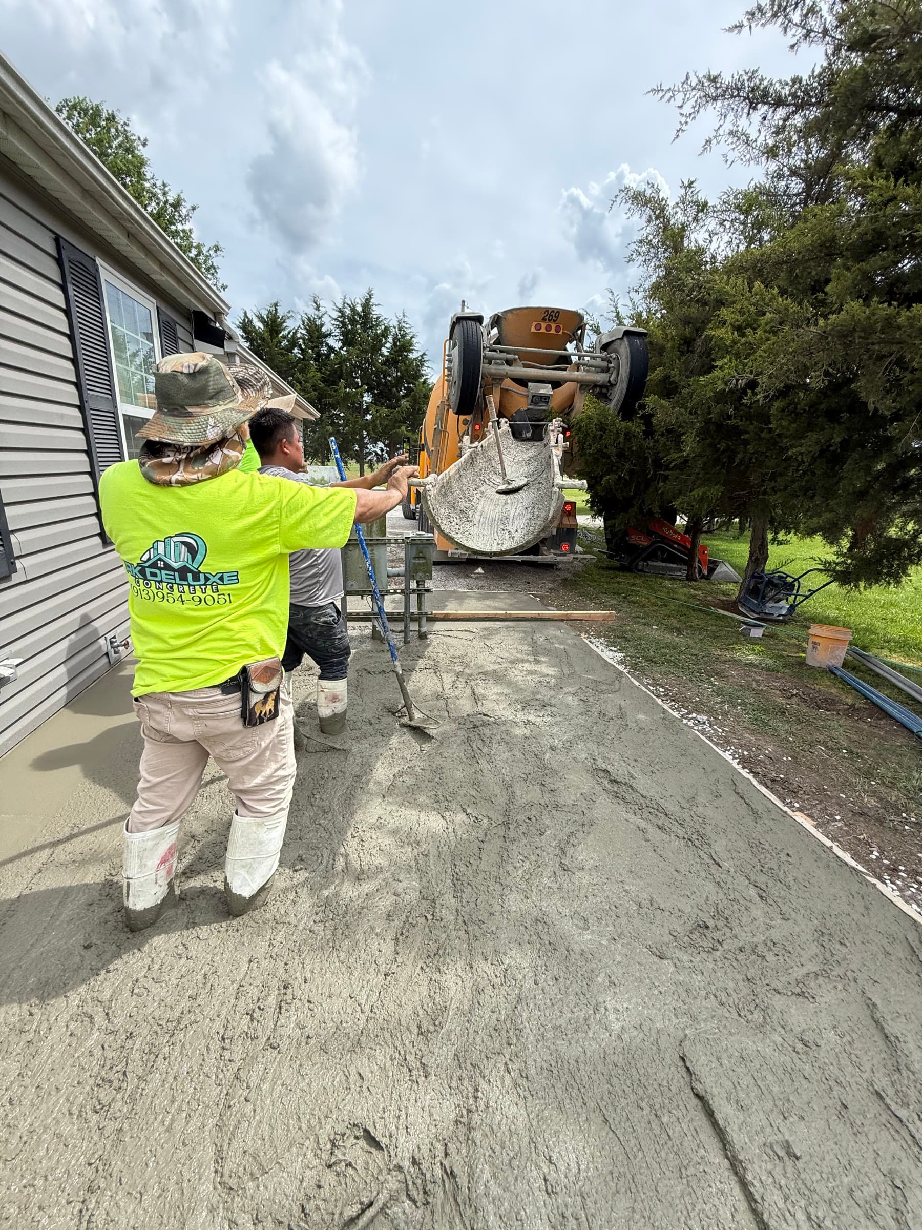 RK Deluxe Concrete crew member in branded work shirt guiding the concrete chute during a pour, mixer truck in background