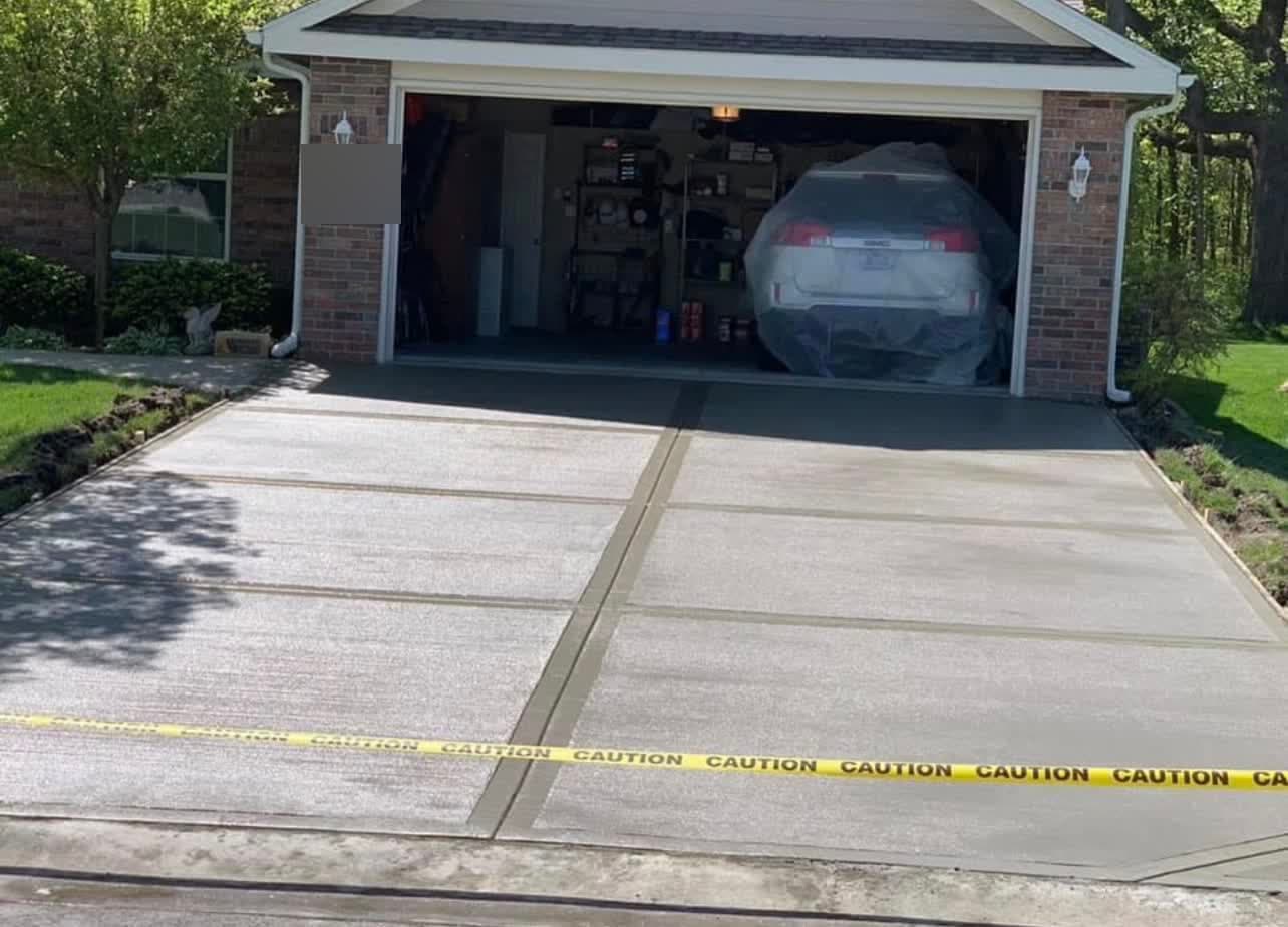 New concrete driveway leading straight to a two-car garage at a brick home, mature trees on either side