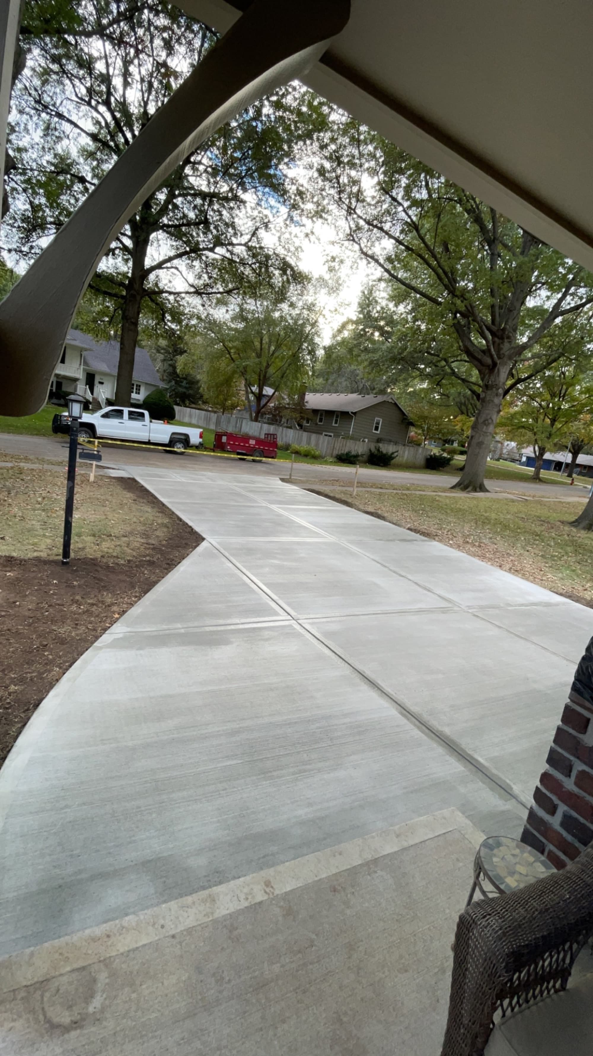 View from a covered porch down a finished concrete walkway toward the street, framed by autumn trees.